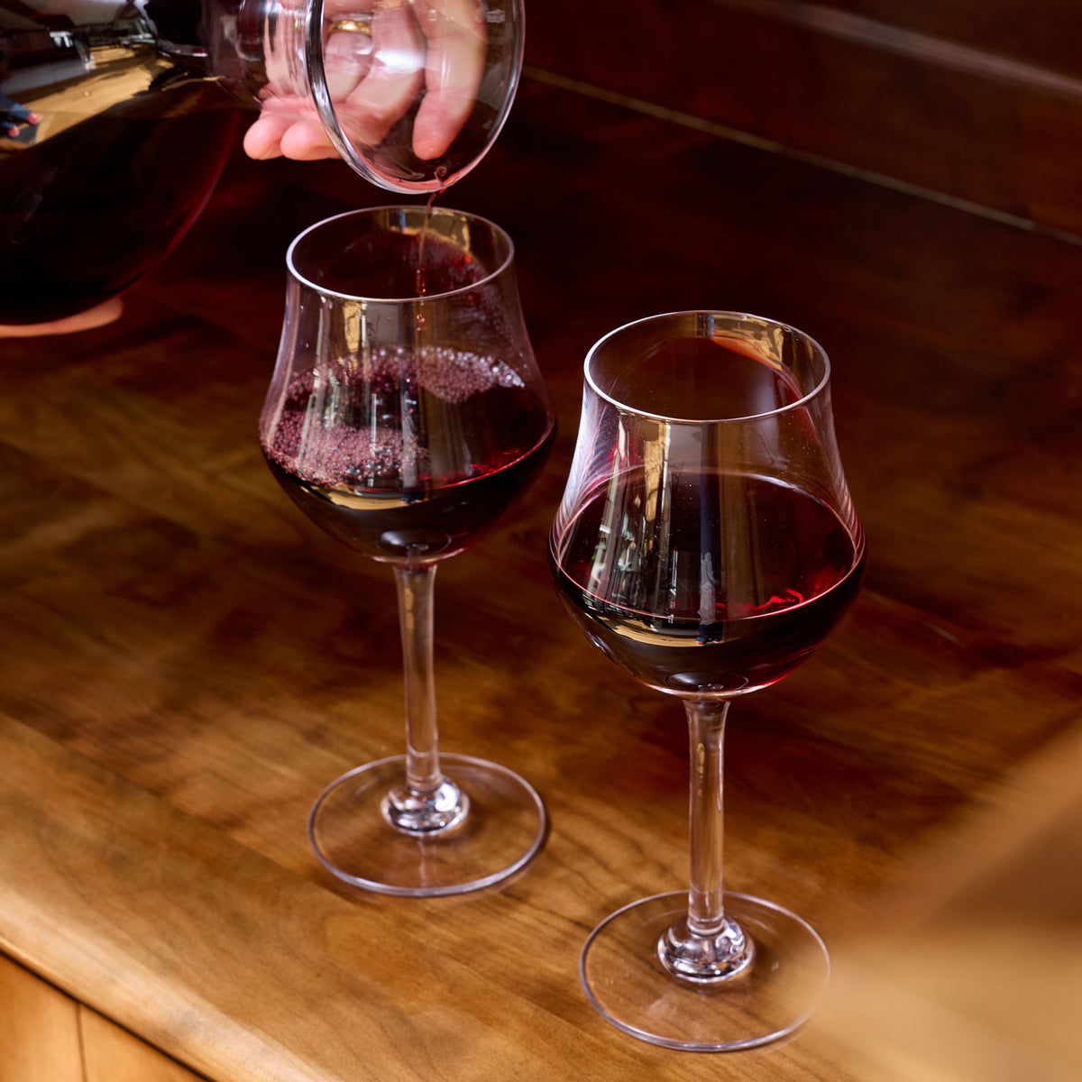 Two clear wine glasses filled with red wine, one being poured from a bottle, on a wooden surface.