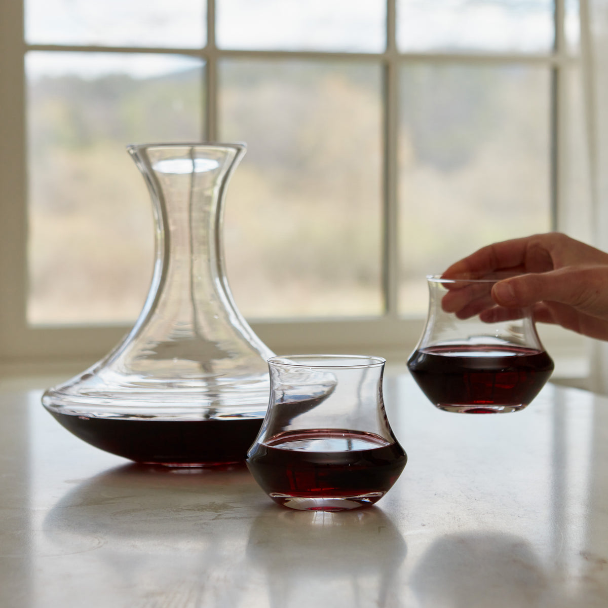 Glass decanter and two stemless glasses containing red wine on a white surface with a hand holding one glass.