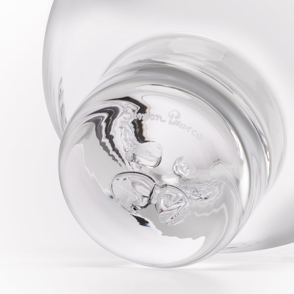 A close-up studio shot of a clear glass object, showing a rounded transparent body and a reflective base with the faint embossed brand name 'Simon Pearce'.