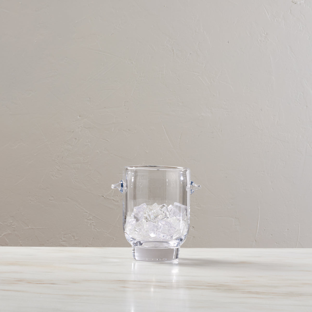 Clear glass ice bucket with two handles, filled with ice cubes.