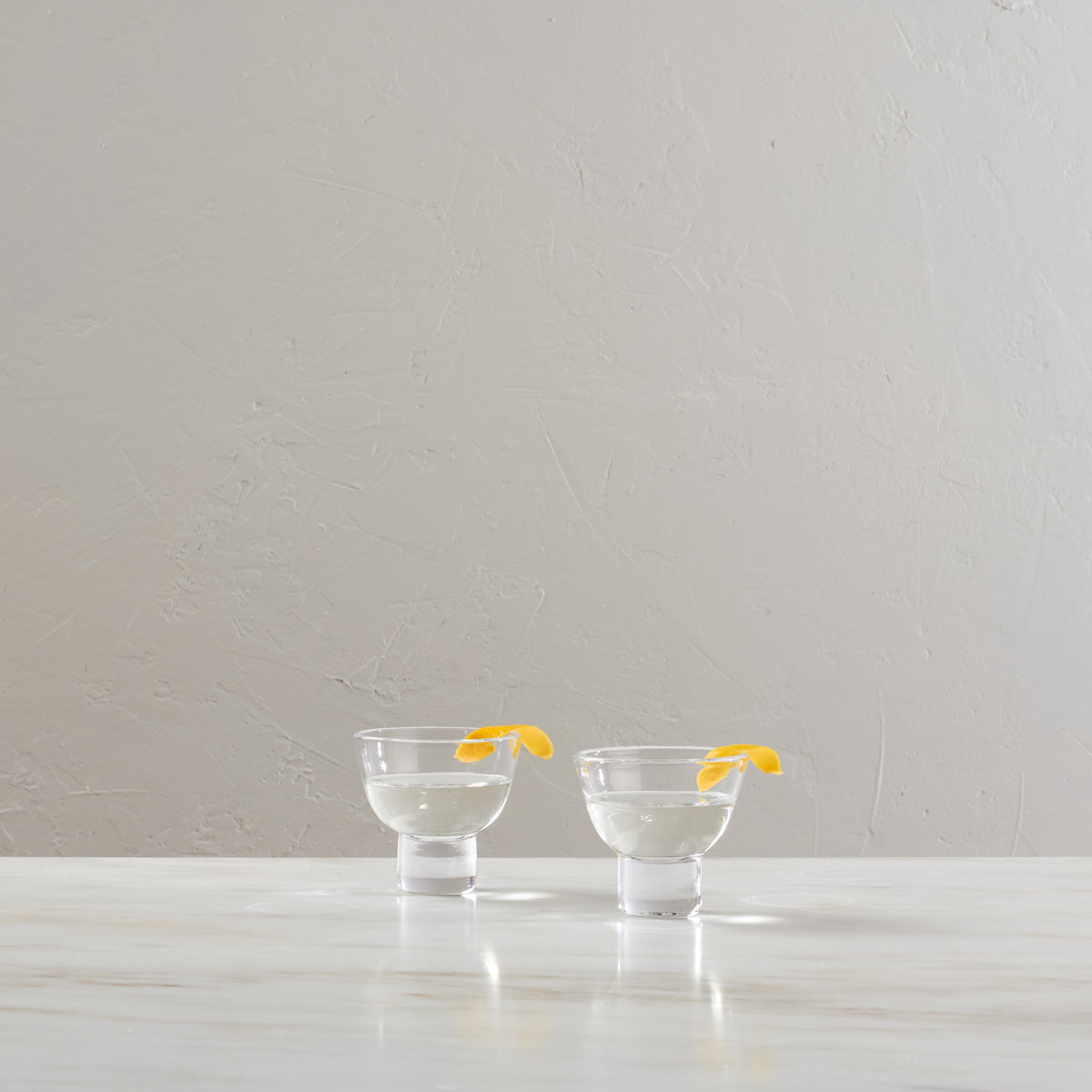 Two clear martini glasses filled with clear liquid and yellow lemon twists, displayed on a white surface.
