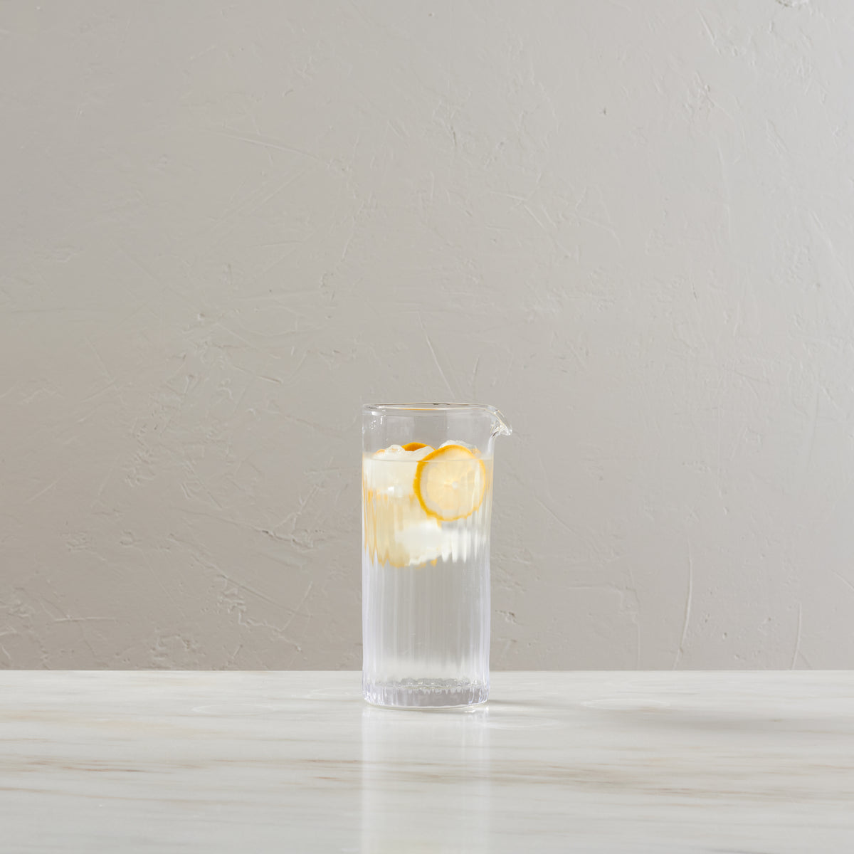 Tall clear glass pitcher with vertical ribbed texture, filled with water, ice, and lemon slices.