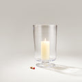 Clear glass hurricane vase with pedestal base holding a lit cream-colored pillar candle.