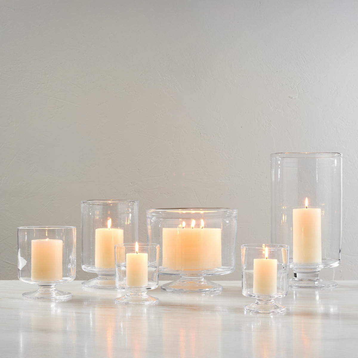 Set of seven clear glass hurricane candle holders in varying heights with lit cream-colored candles inside.