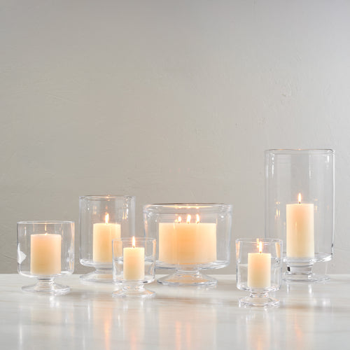 Set of seven clear glass hurricane candle holders in varying heights with lit cream-colored candles inside.