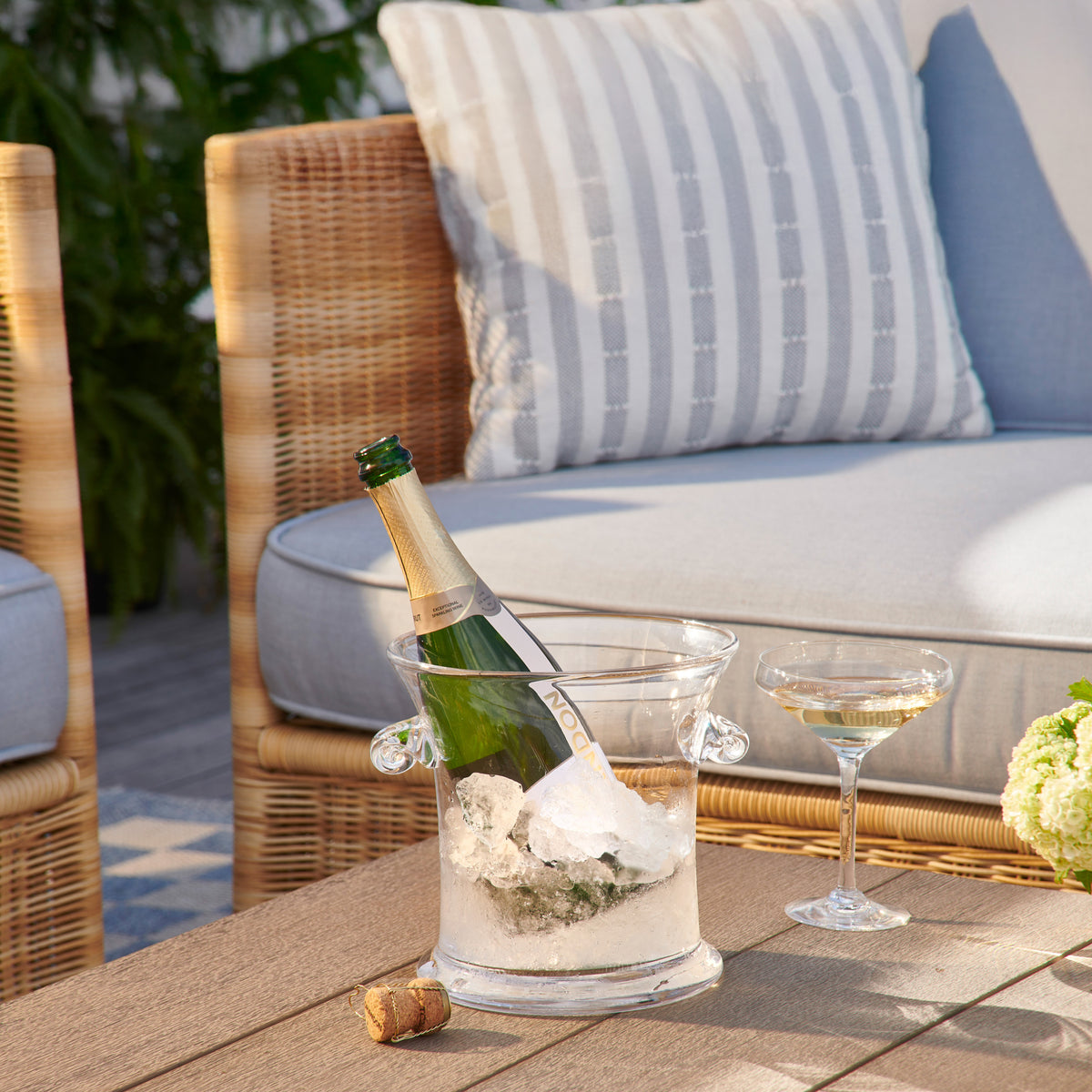Clear acrylic ice bucket holding a champagne bottle surrounded by ice, displayed on outdoor table.