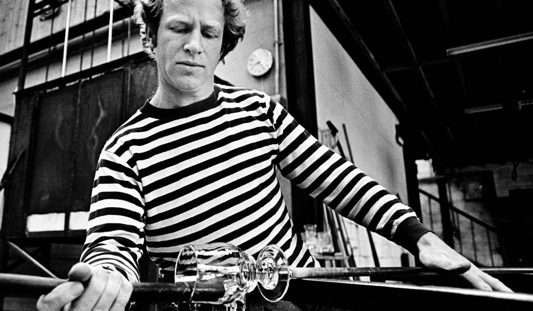 Black and white photograph of a young Simon Pearce in a striped shirt shaping a handblown glass piece on a metal rod at his glassblowing bench in his studio