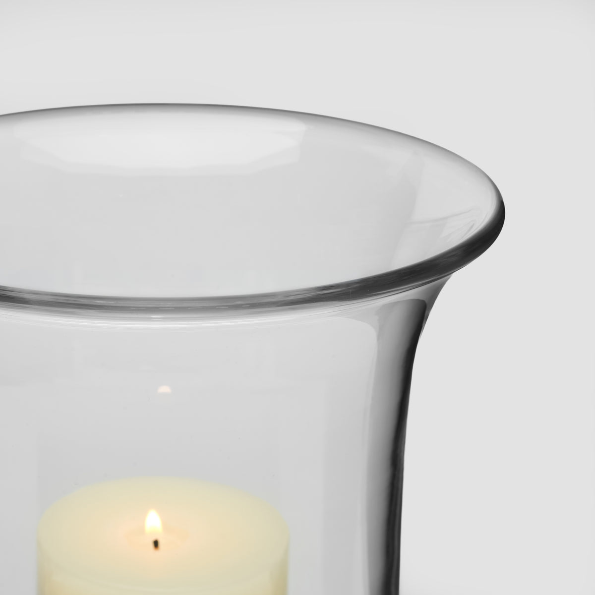 Clear glass hurricane with flared rim containing a lit cream-colored candle.