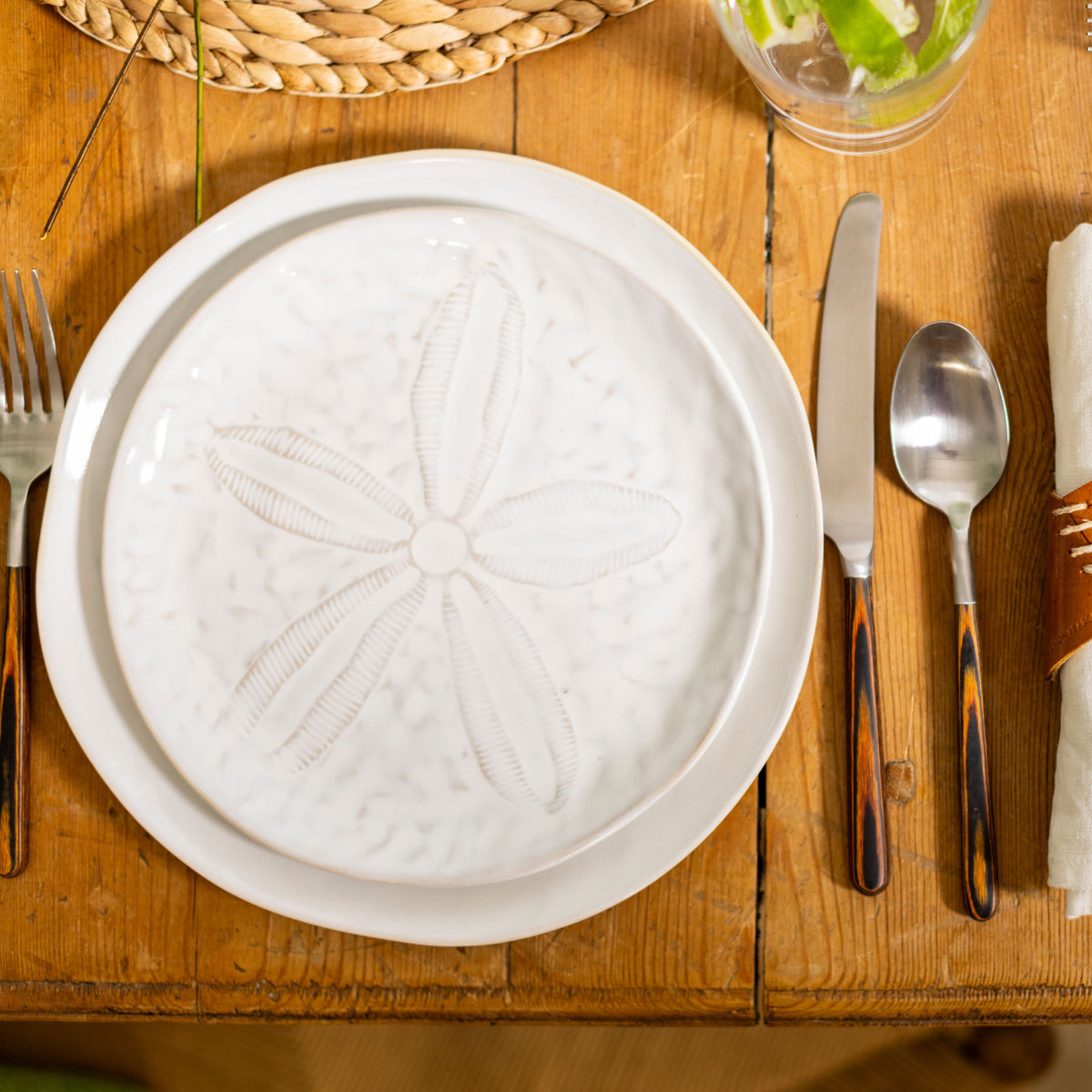 White side plate with embossed sand dollar design, set on wooden table with silverware.