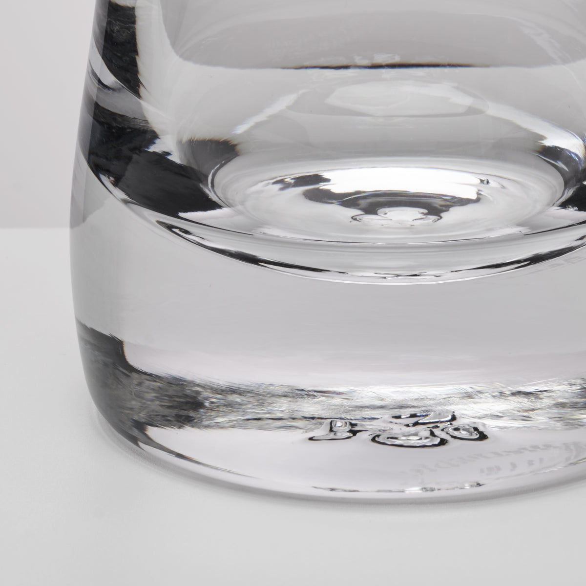 Close-up of a clear glass showing the base and lower body with reflective glass surfaces on a light gray background.
