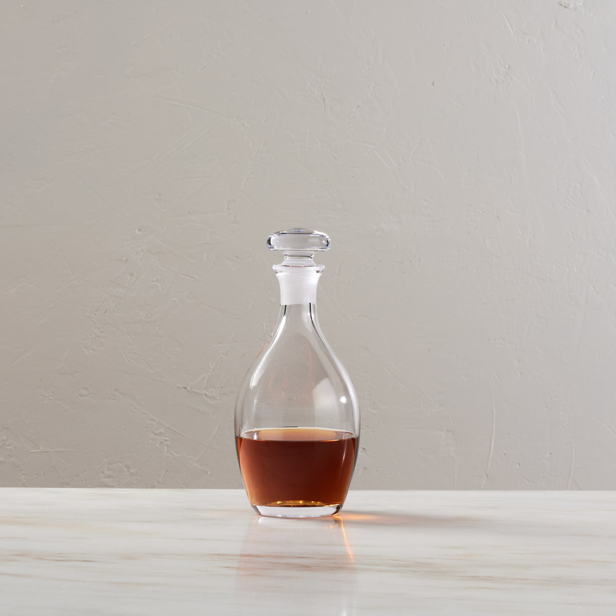 Clear glass decanter with stopper containing amber liquid, on white surface against neutral background.