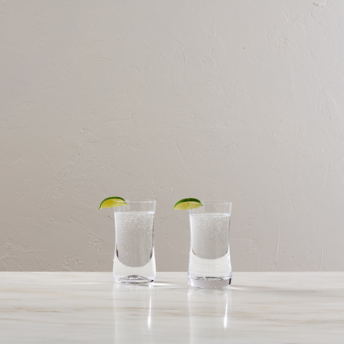 Two clear glasses filled with sparkling water and lime slices on white surface.