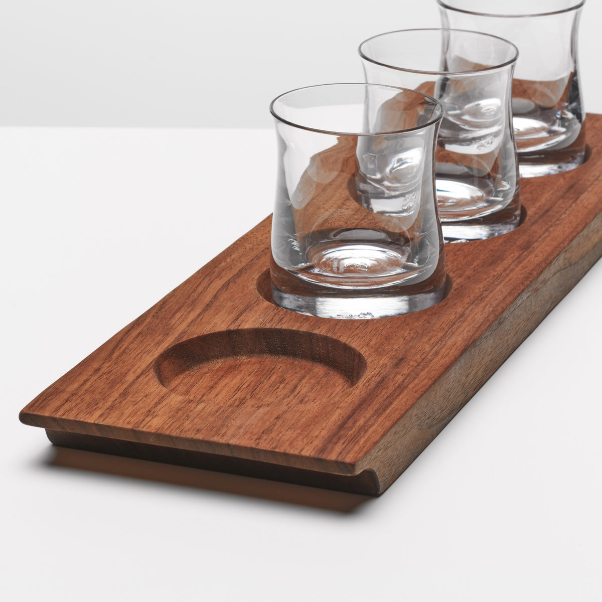 Three clear glass whiskey glasses on walnut wood base with circular bottle holder.