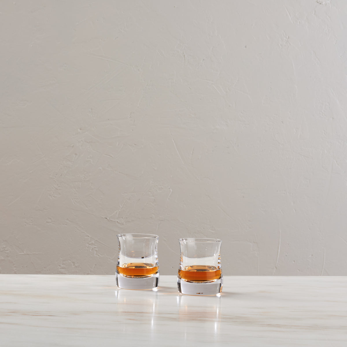 Two clear whiskey glasses filled with amber liquid on a white marble surface against a textured beige wall.