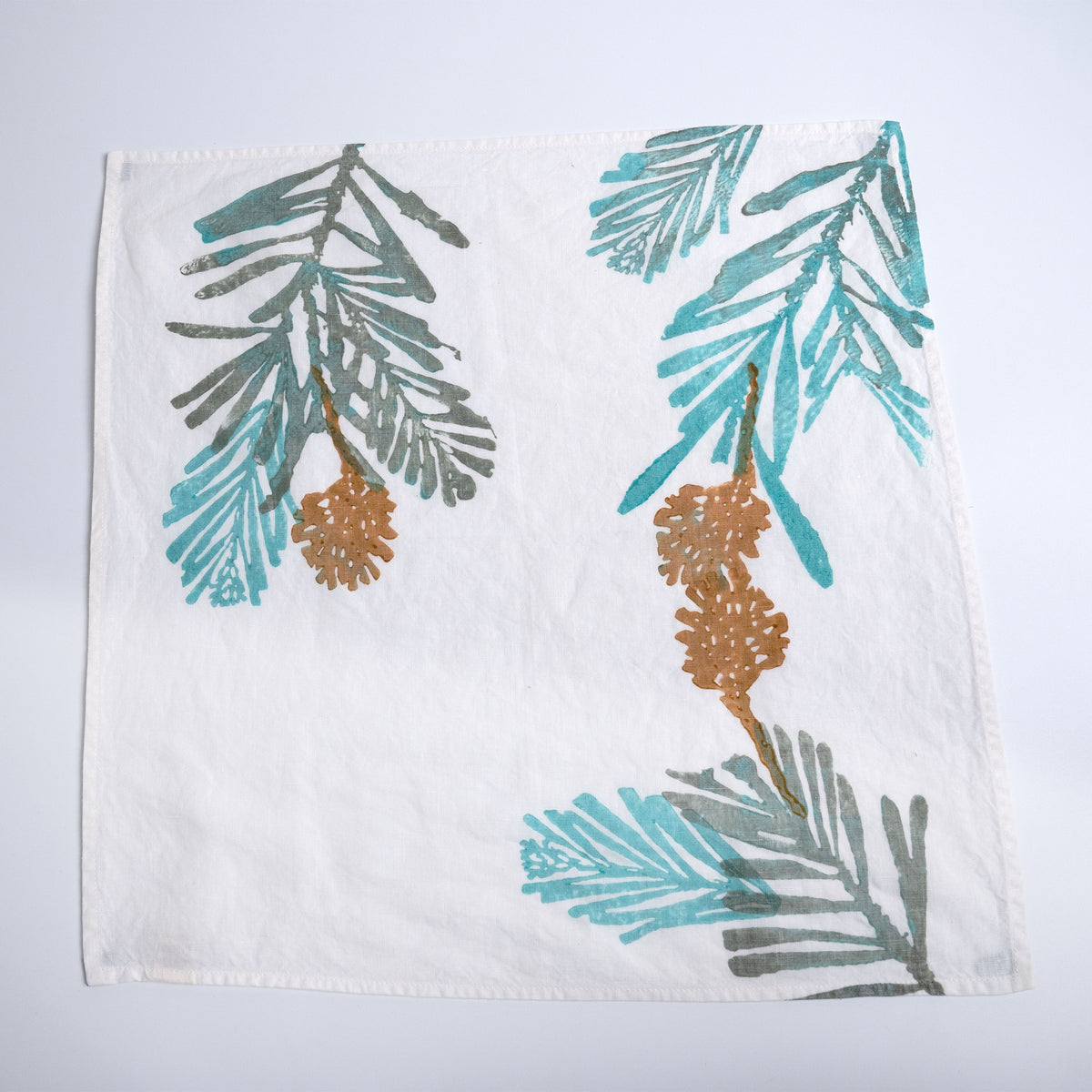 White linen napkin with pine branch and pinecone design in teal, gray, and gold.
