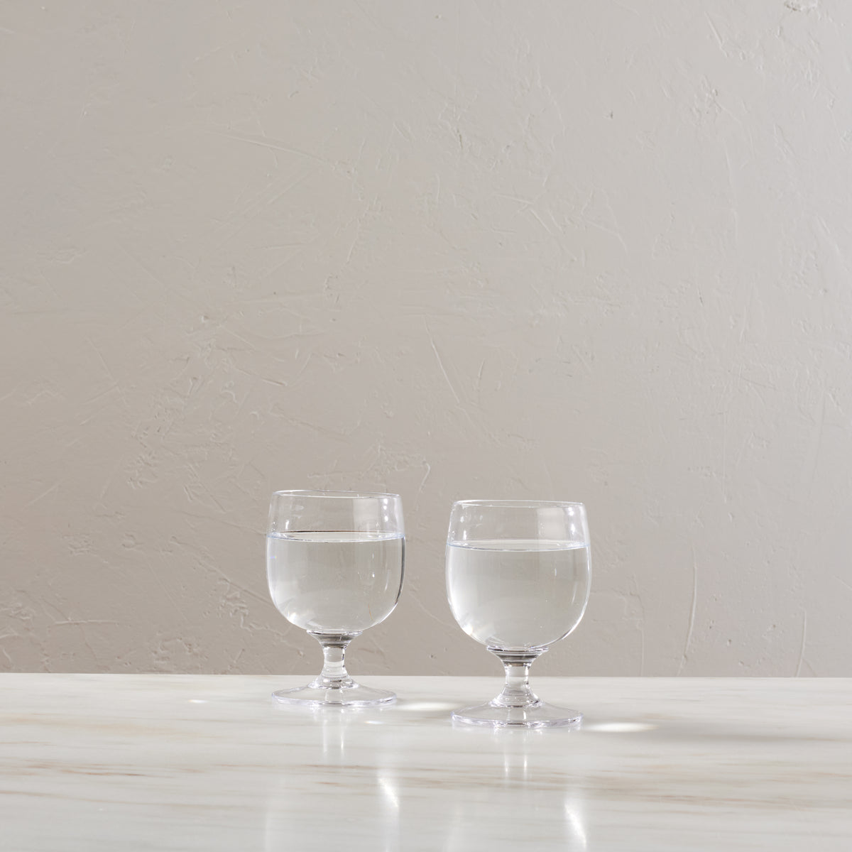 Two clear glass goblets with pedestal bases filled with water, displayed on a white surface.