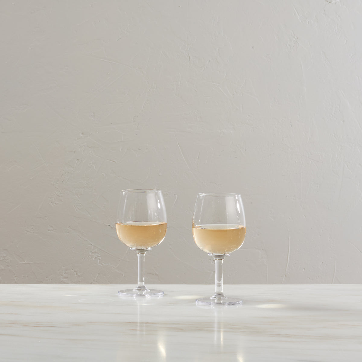Two clear wine glasses filled with white wine on a white surface against a neutral background.