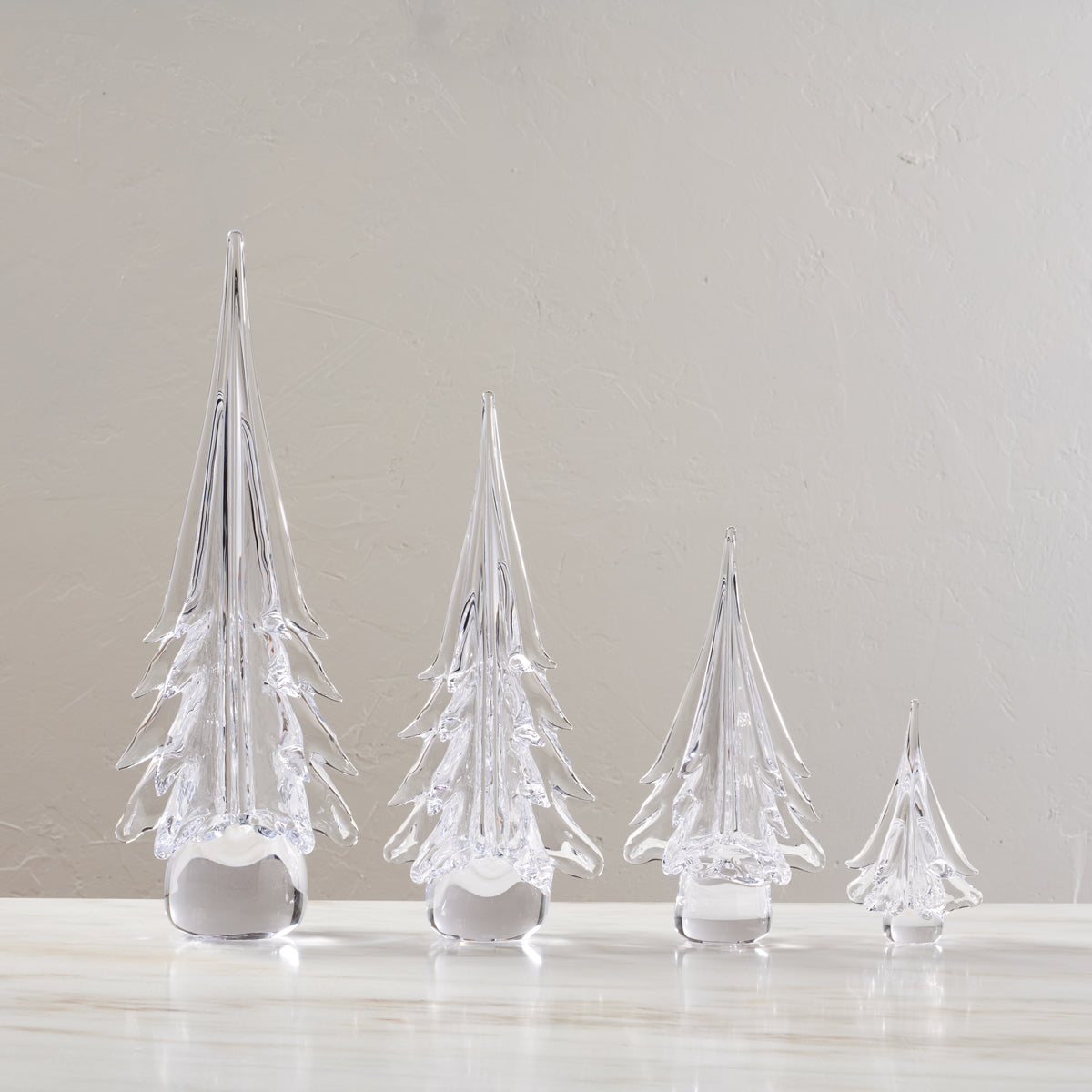 Four clear glass evergreen tree-shaped decorations in decreasing sizes with white frosted bases.