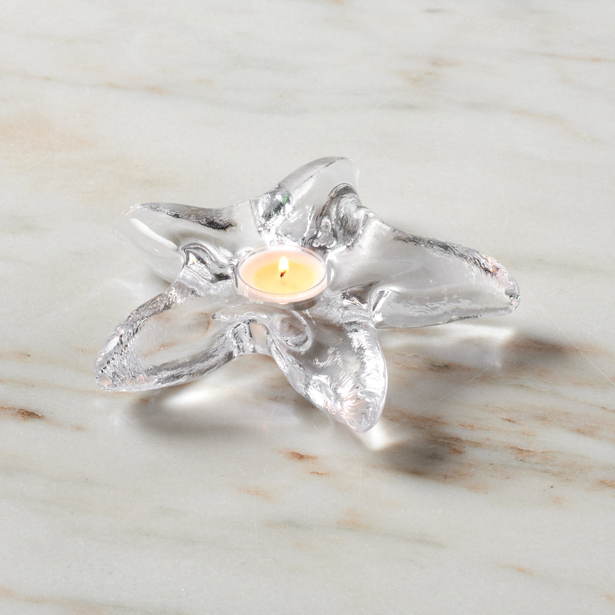 Clear glass starfish-shaped tealight holder with lit cream candle in center on marble surface.