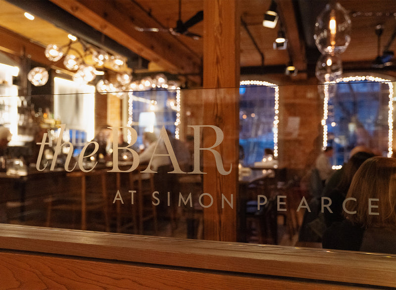 The Bar at Simon Pearce sign etched in glass, with warm amber lighting and the rustic wood interior visible behind it