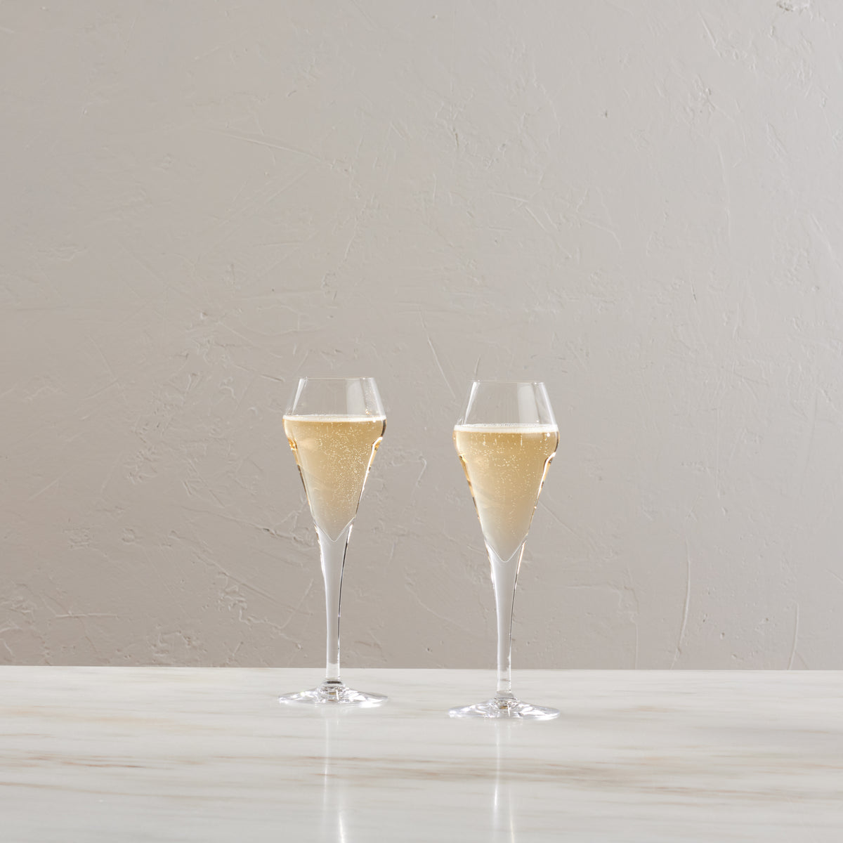 Two clear flute glasses filled with sparkling wine on a white surface.