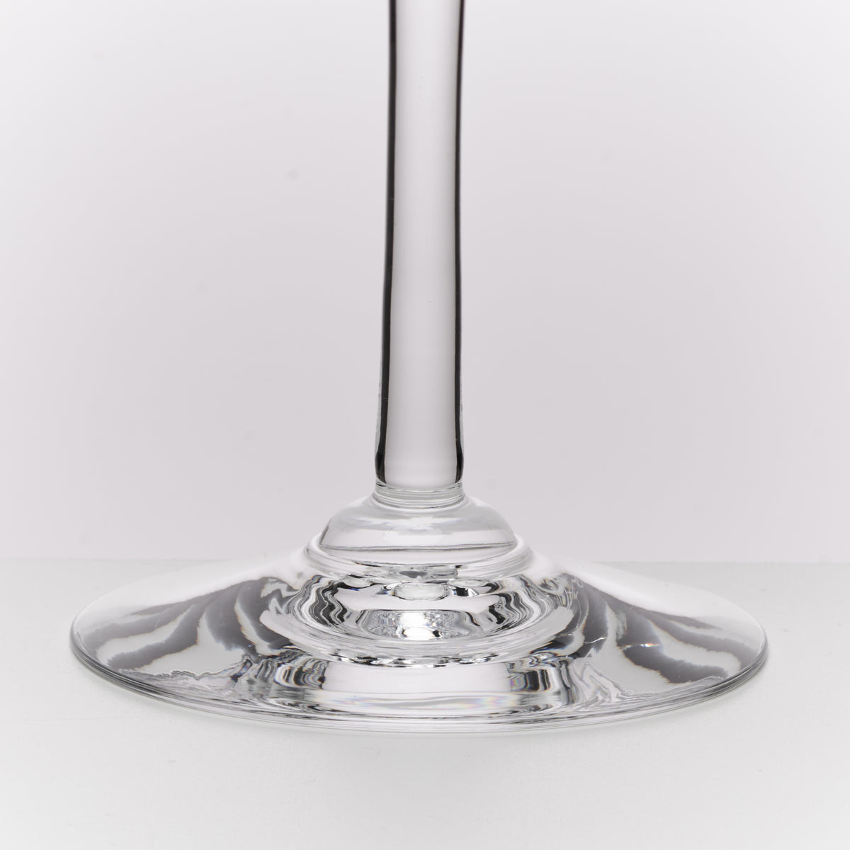 A close-up studio shot of a clear glass wine glass stem and wide circular base on a light gray background.