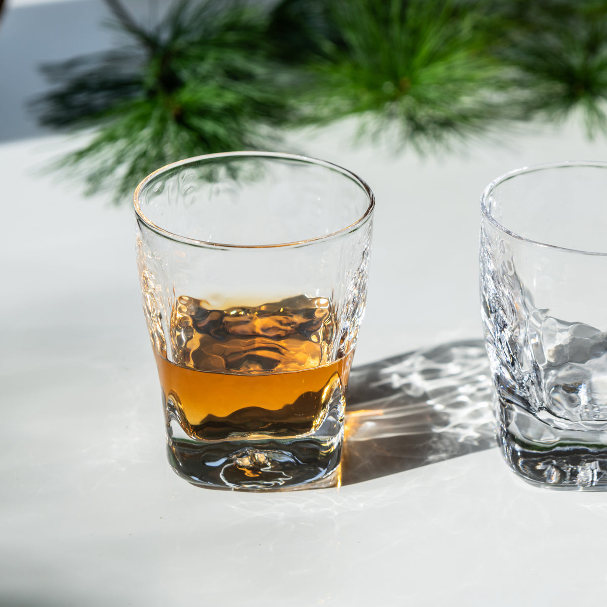 Two crystal rocks glasses, one filled with amber bourbon and one empty, with gold-rimmed details and pine branches in backgro