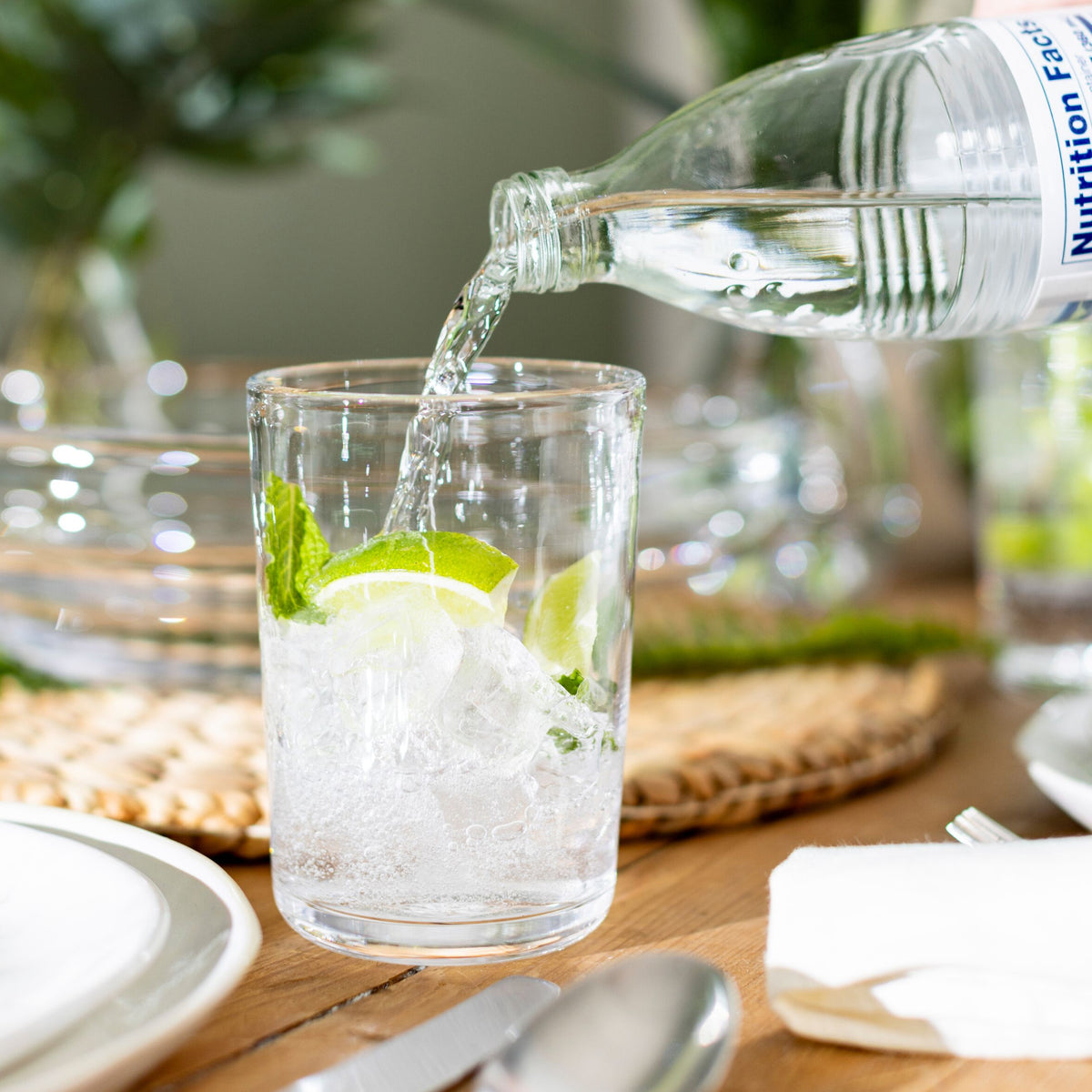 Water pouring into a clear glass containing ice and lime slice on a wooden table with place settings.