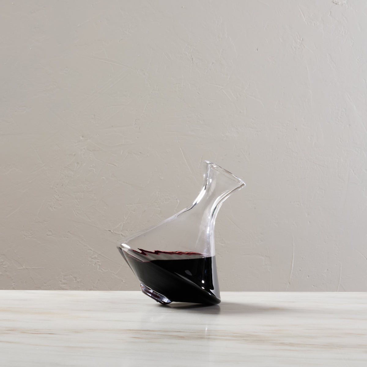 Clear glass wine decanter with curved spout, tilted base, partially filled with red wine.
