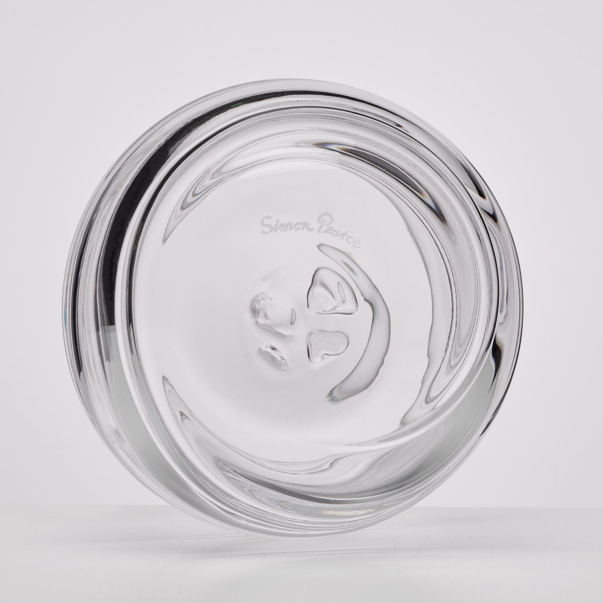 A clear glass ashtray with a sculptural, wavy design and the etched brand name Simon Pearce visible in the center, shown on a light gray background.