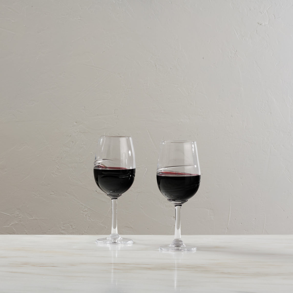 Two glasses filled with red wine on a white surface against a light gray textured wall.