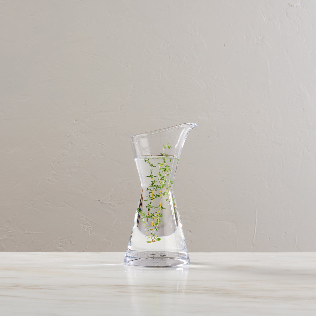 Clear glass vase with hourglass shape and flared rim, filled with water and small green plant sprigs.