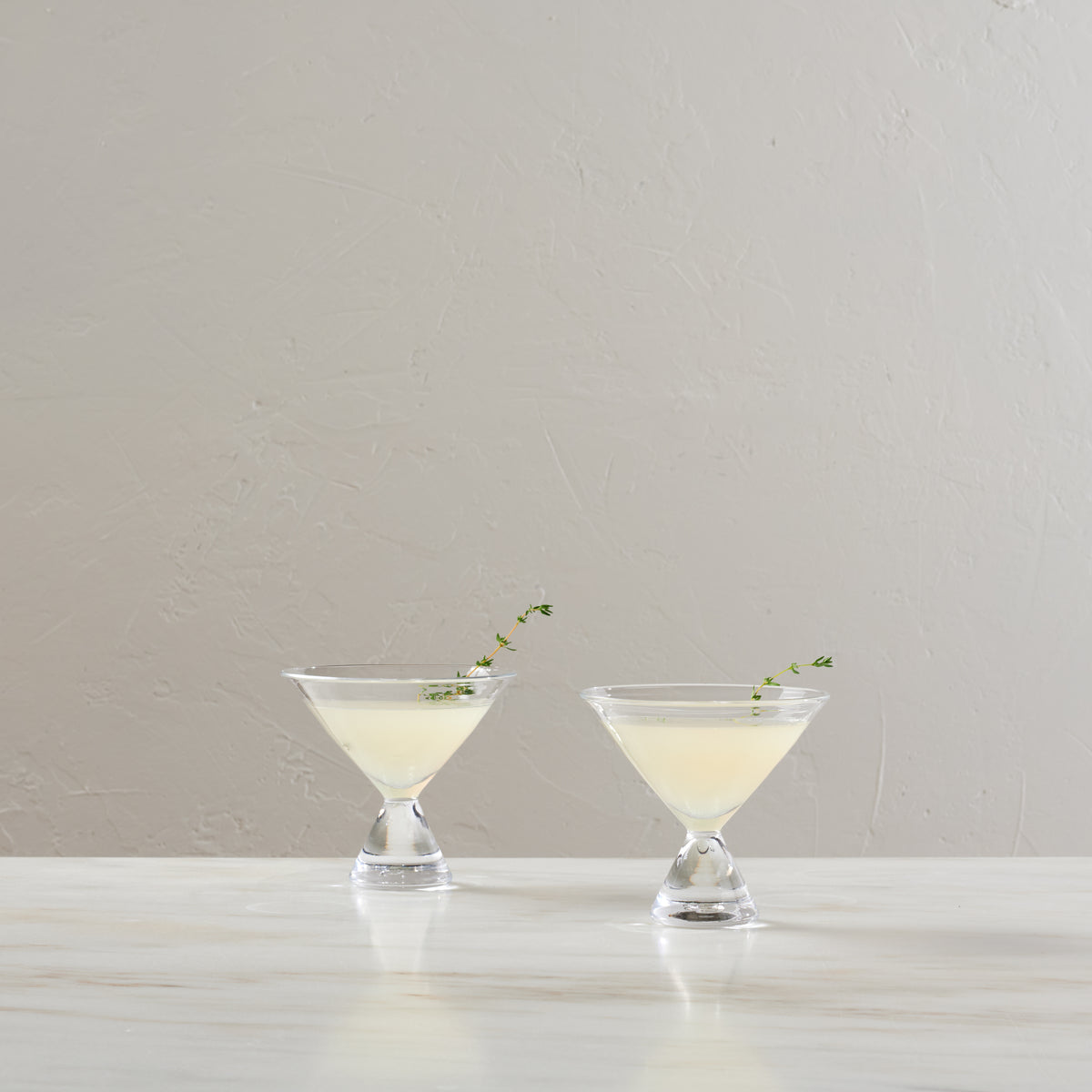 Two clear stemless martini glasses filled with pale cocktail, each garnished with herbs.