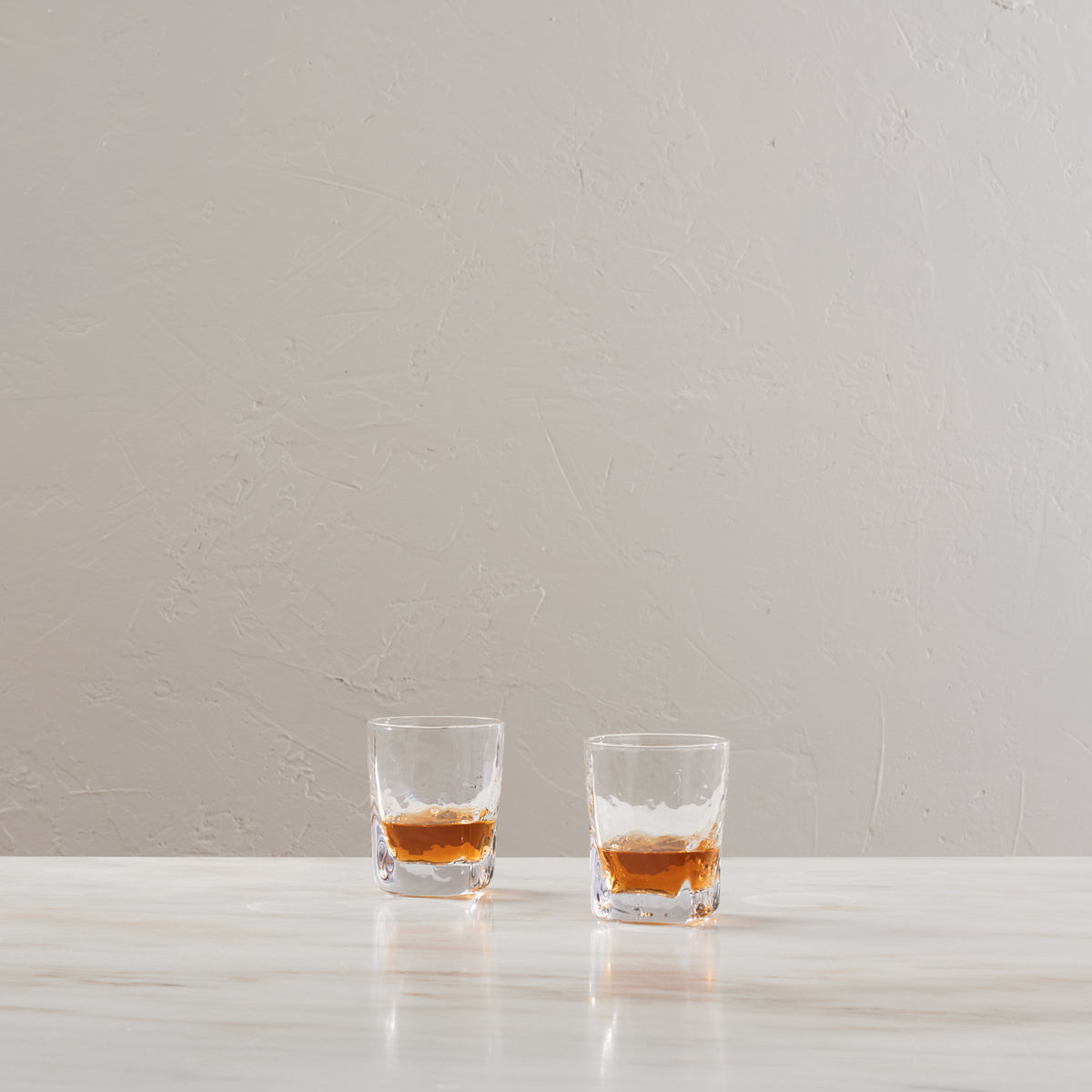 Two glasses filled with bourbon whiskey on a white surface against a textured beige wall.