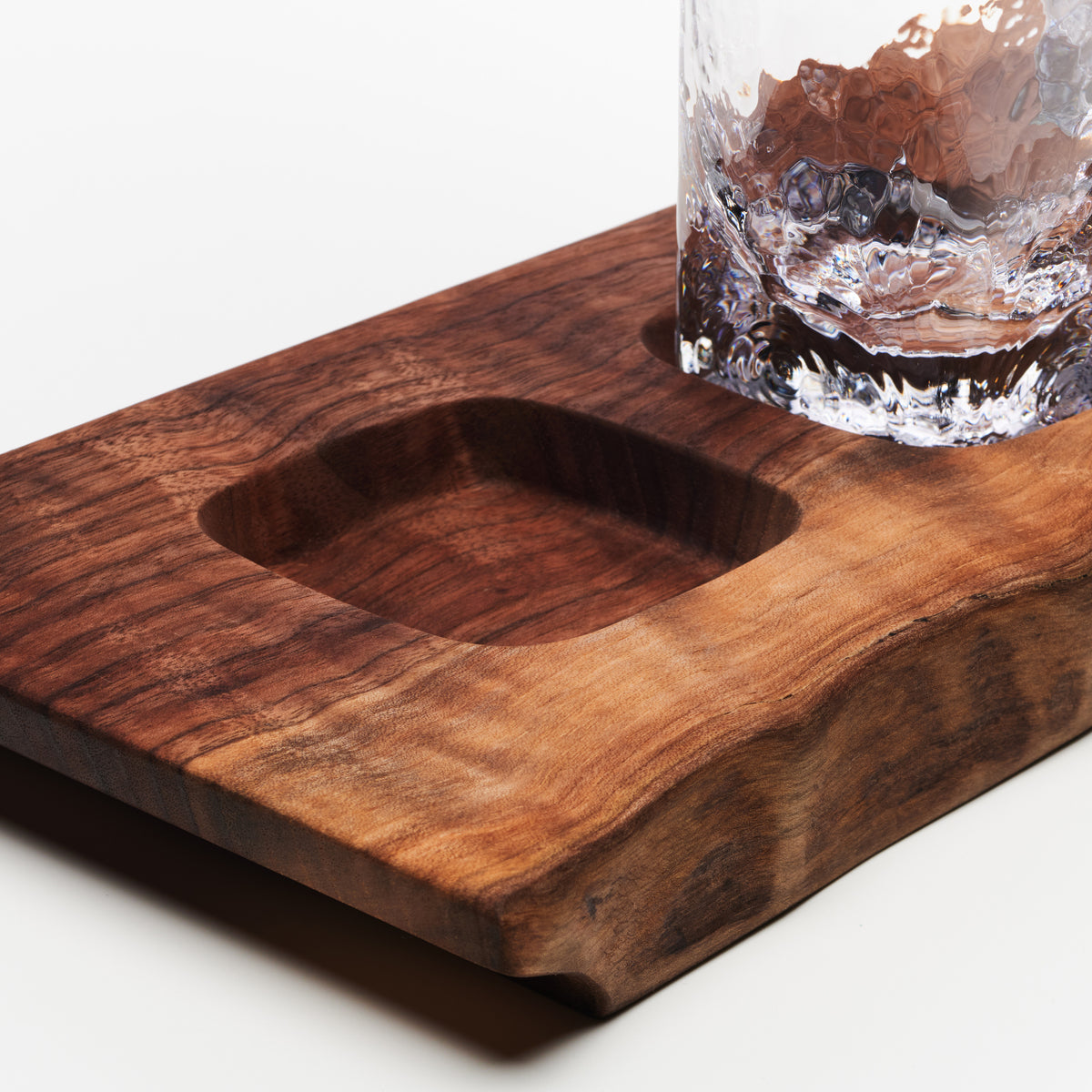 Wooden serving board with carved recessed sections holding a rocks glass filled with bourbon and ice.