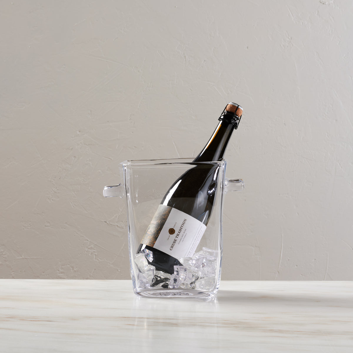 Clear glass ice bucket with two handles holding a champagne bottle filled with ice cubes.