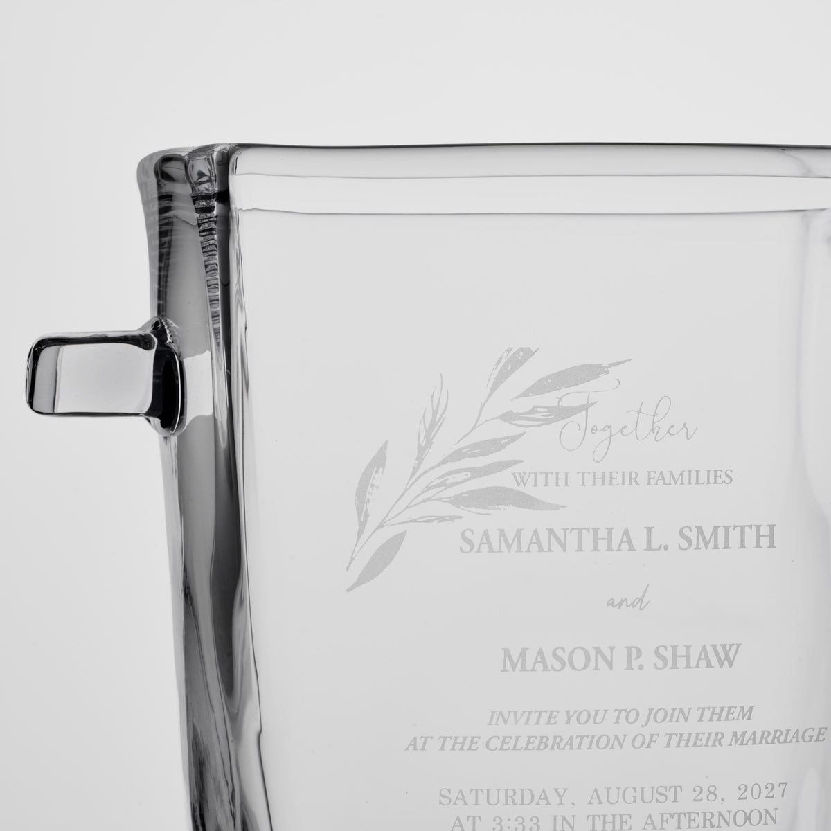 Clear glass ice bucket with black handle featuring engraved wedding invitation text and botanical leaf design.