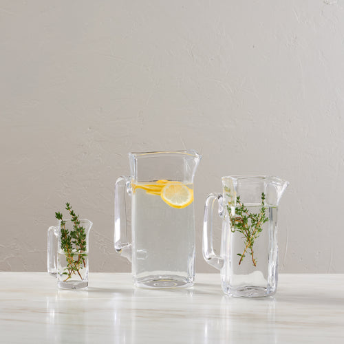 Clear glass pitcher with handle, containing water and lemon slice, displayed with two smaller matching glasses holding sprigs