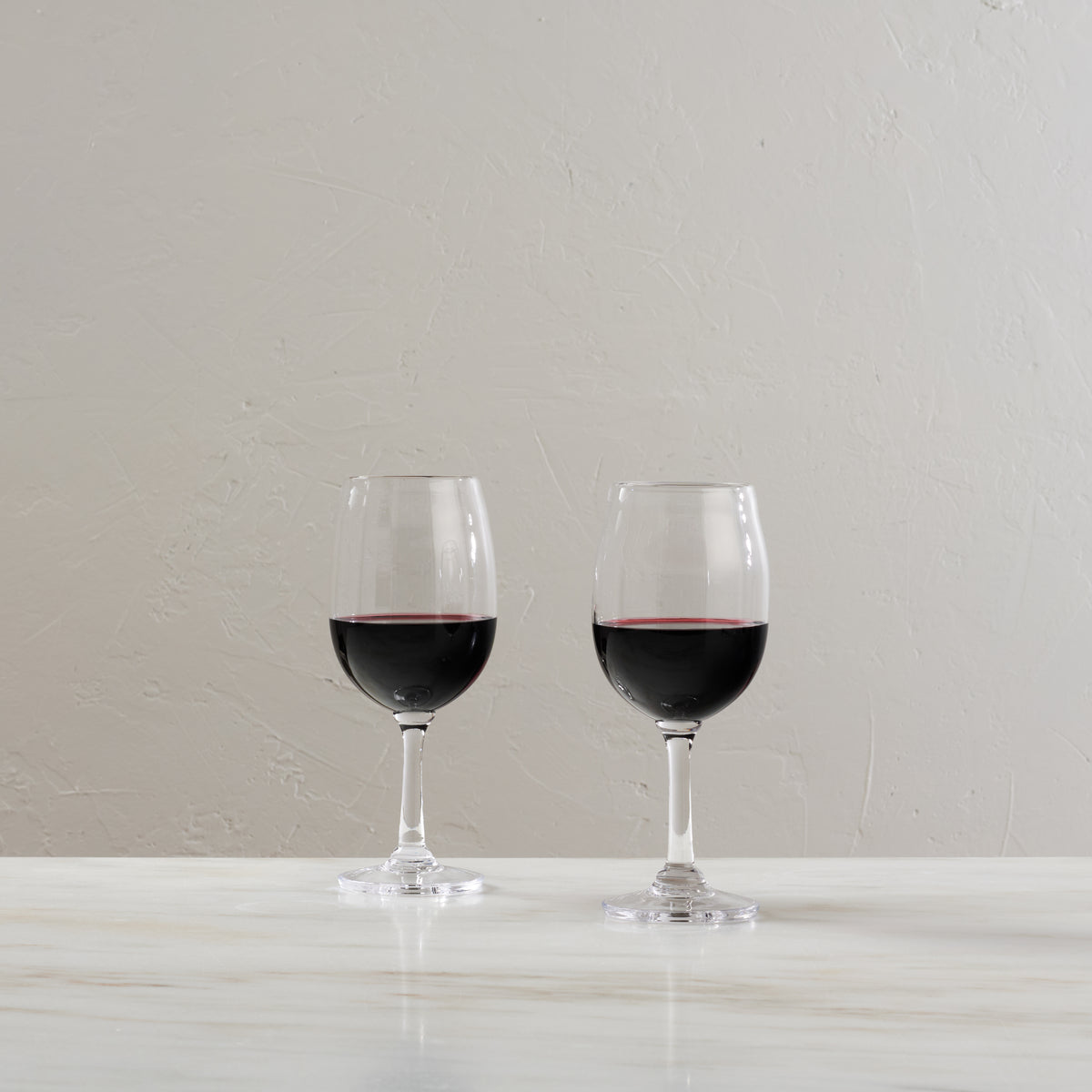 Two glasses filled with red wine on a white surface against a light textured wall.
