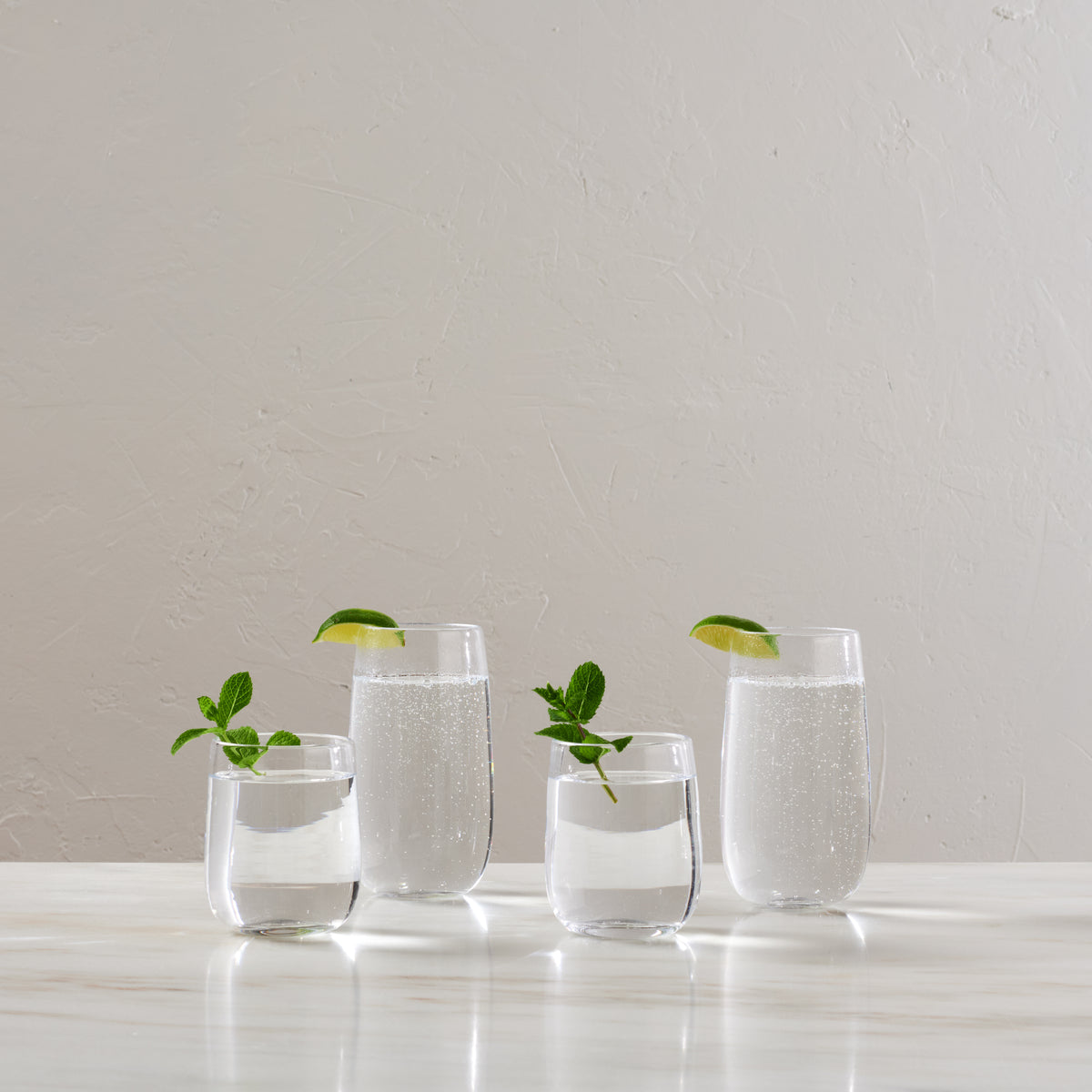 Four clear glass tumblers with lime slices and mint sprigs, two filled with clear liquid and two empty.
