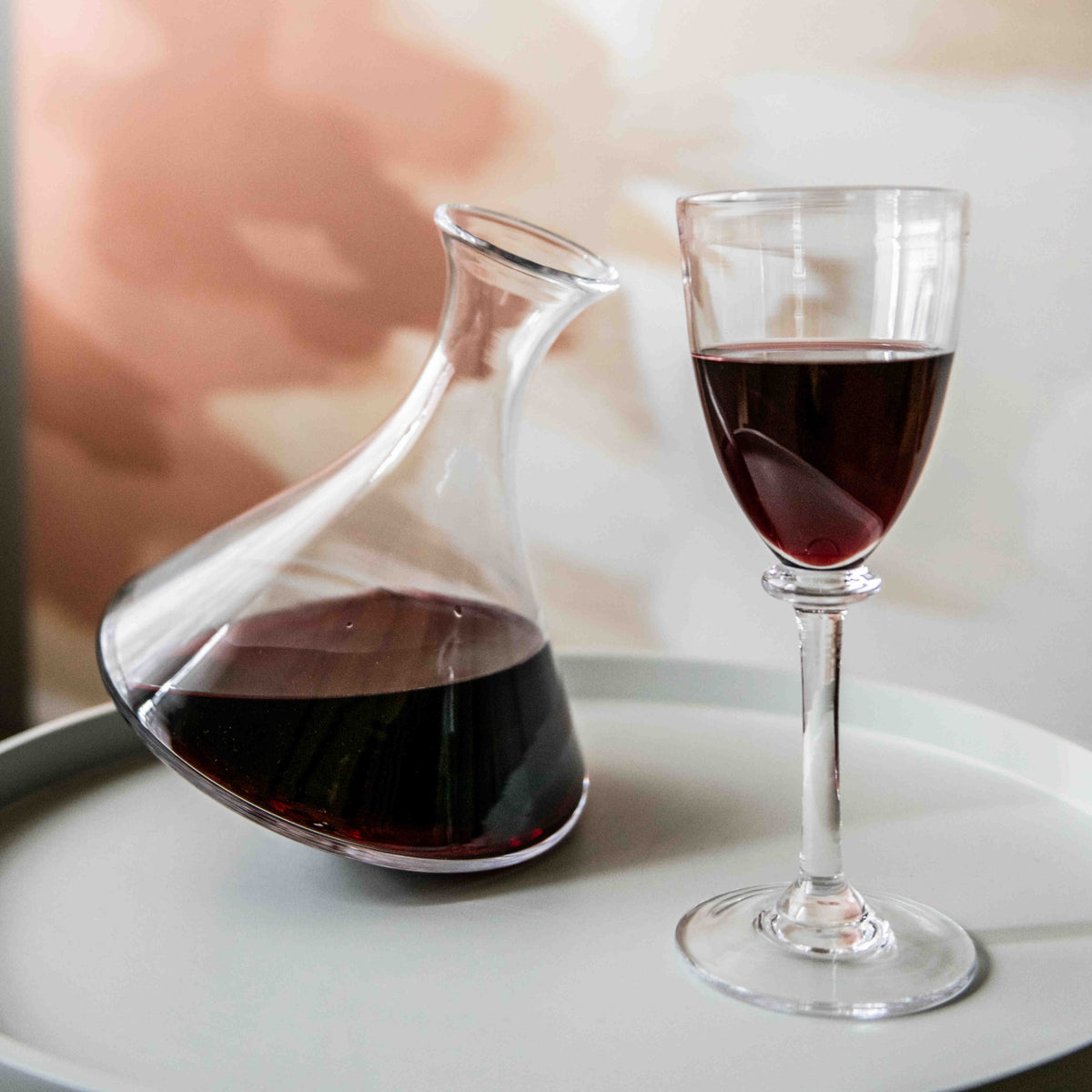 Glass wine decanter with curved spout filled with red wine, displayed next to a filled wine glass on a white plate.