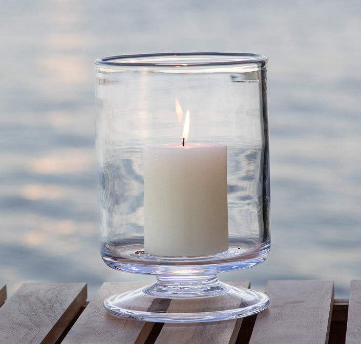 A glass Nantucket Hurricane with a lit pillar candle, sitting on a wooden dock with a lake in the background