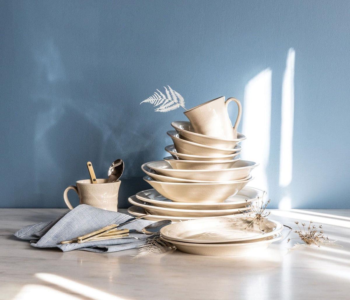 Stacked ivory dinnerware with crackle texture, including plates, bowls, and a mug with handle, arranged with utensils and nap