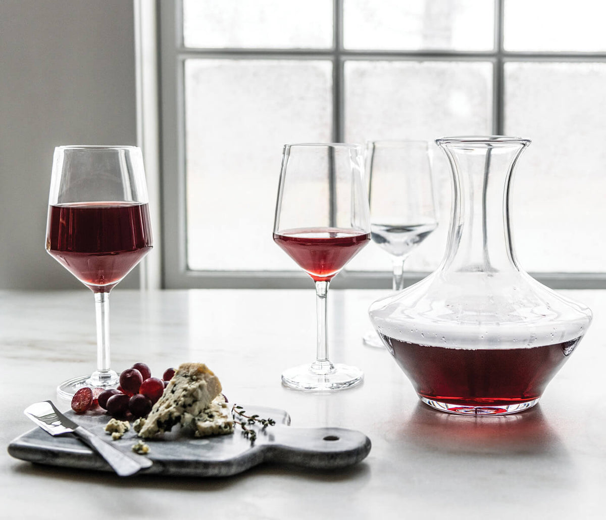 Clear glass decanter filled with red wine, displayed with two filled wine glasses and empty glass, next to cheese and grapes 