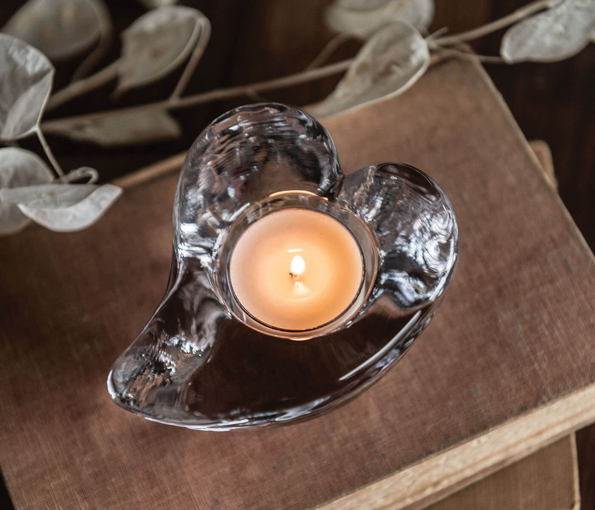 Heart-shaped clear glass tealight holder with lit cream-colored candle on tan fabric surface.