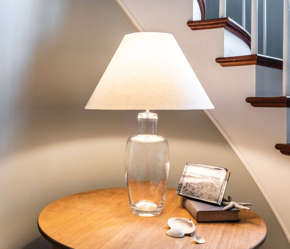 Tall table lamp with clear glass base filled with sand and cream trapezoid shade, lit on wooden surface.