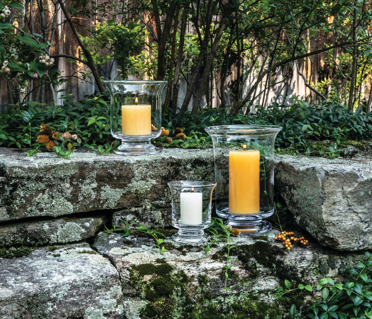 Three glass hurricane candles in varying heights with white and yellow wax, displayed on moss-covered stone outdoors.