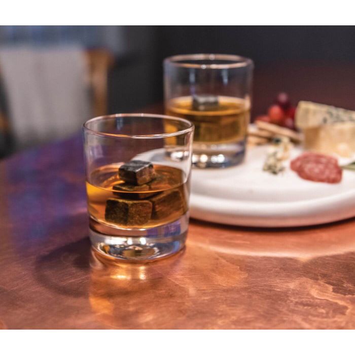 Two whiskey glasses with ice and amber liquid on a wooden table with appetizers and a white plate nearby.