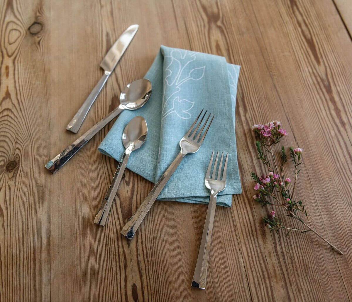 Five-piece flatware set with knife, two spoons, and two forks displayed on blue linen napkin with wood surface.