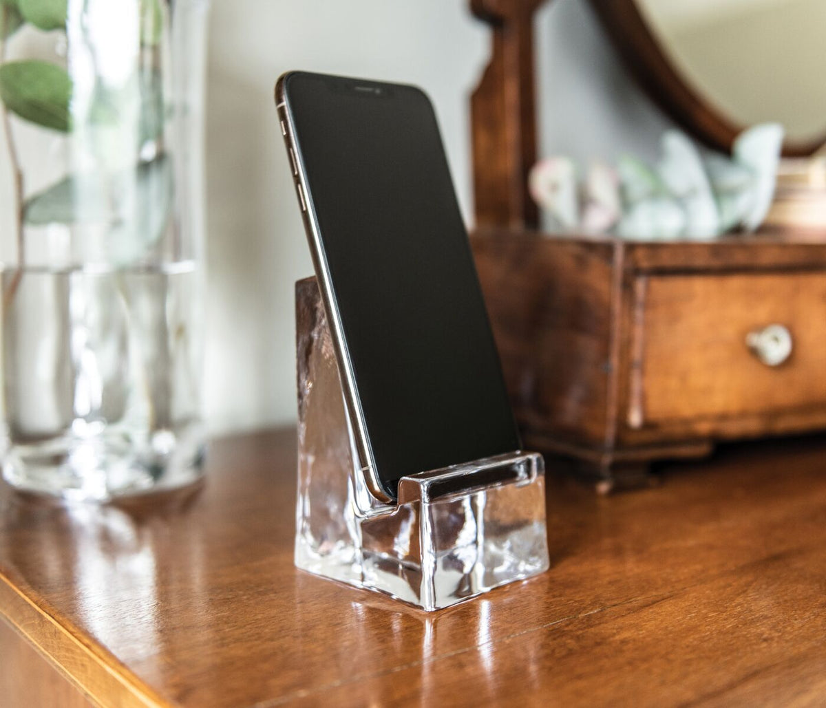 Clear glass phone holder with angled slot holding a smartphone upright on wooden surface.